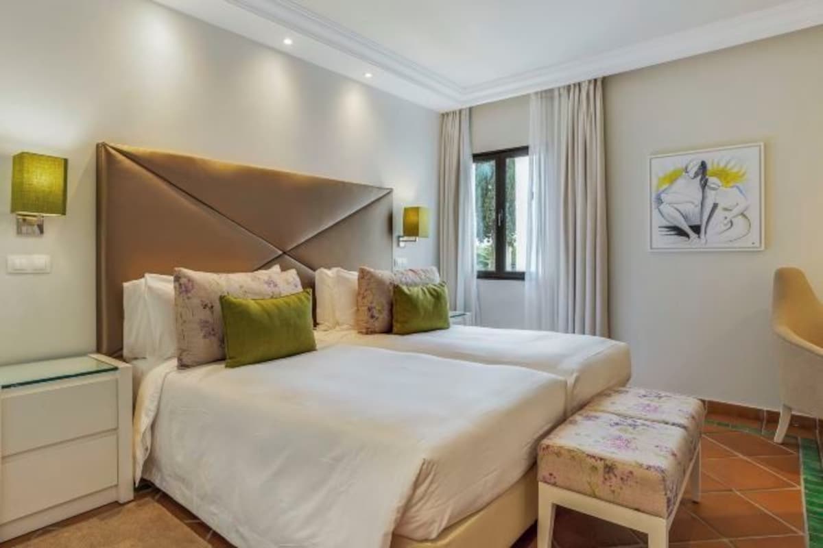 A hotel room with two beds, a bench, a nightstand, and a painting on the wall.