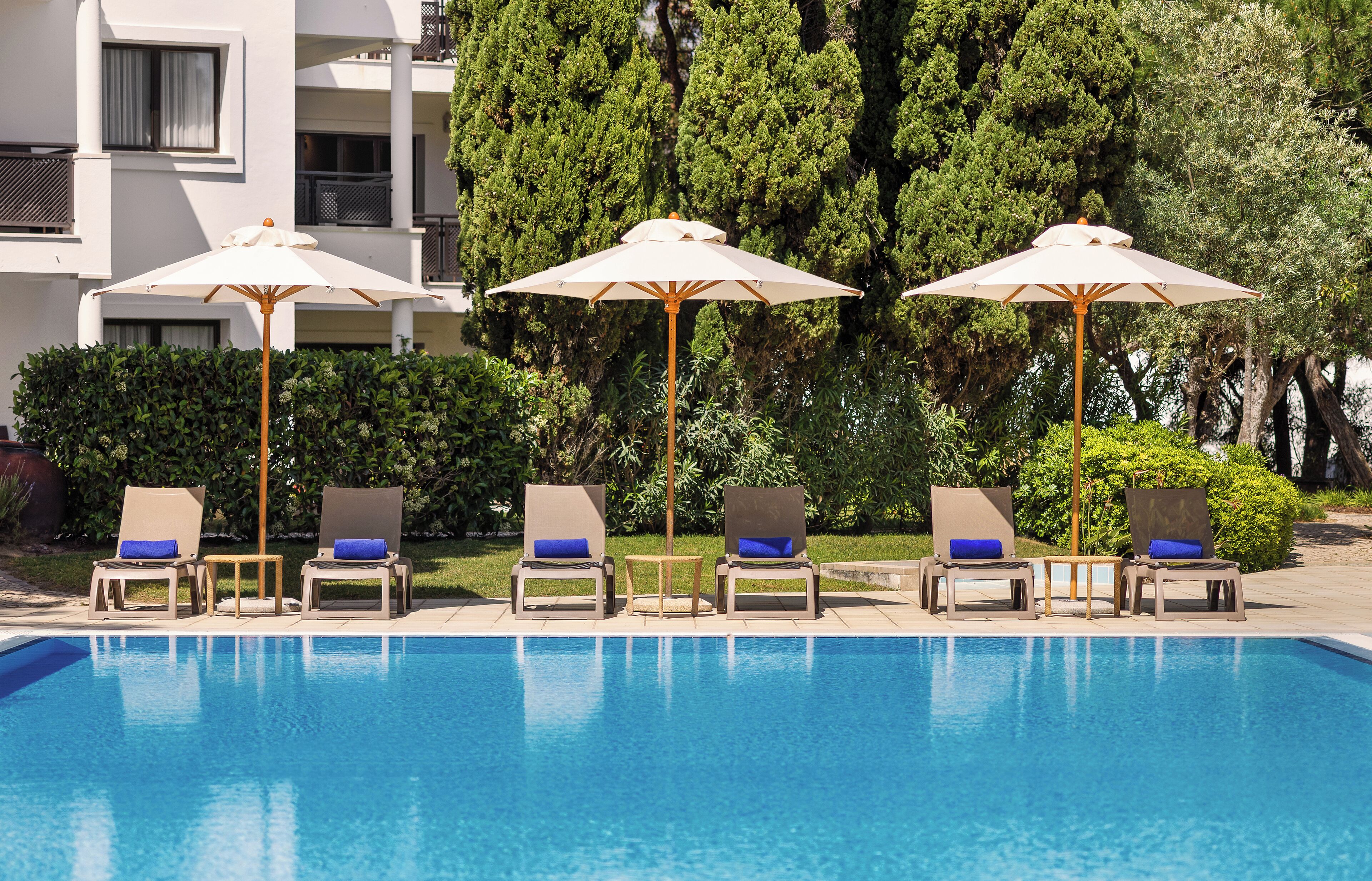 A swimming pool with lounge chairs and umbrellas, surrounded by greenery and a building.