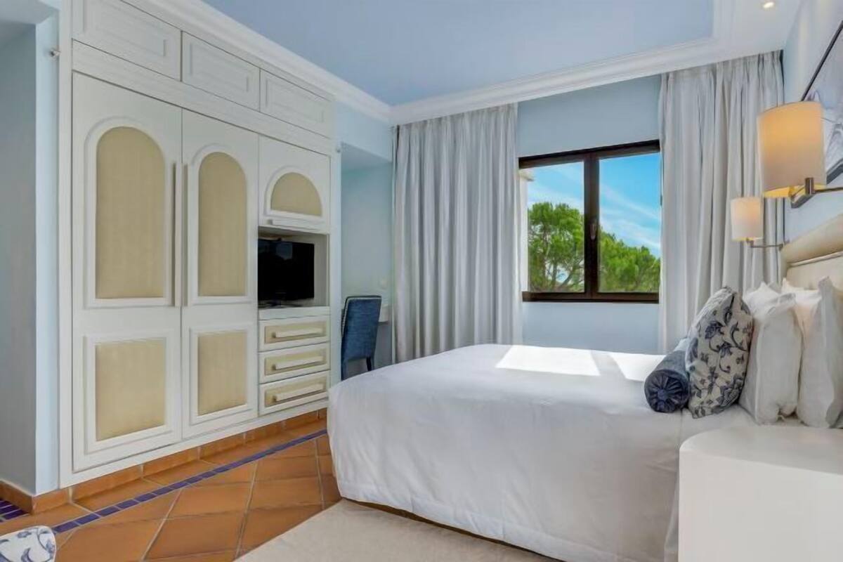 A bedroom with a large bed, a TV, a wardrobe, and a window with curtains.