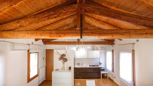 Ruime kamer met een hoog houten plafond, een keukenhoek met moderne apparatuur, en een groot raam dat voor natuurlijk licht zorgt.