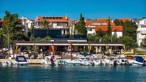 A marina with boats docked, a covered area with seating, and buildings with orange roofs in the background. - Deluxe Villa Laura with Pool Studio 4b (Novalja)