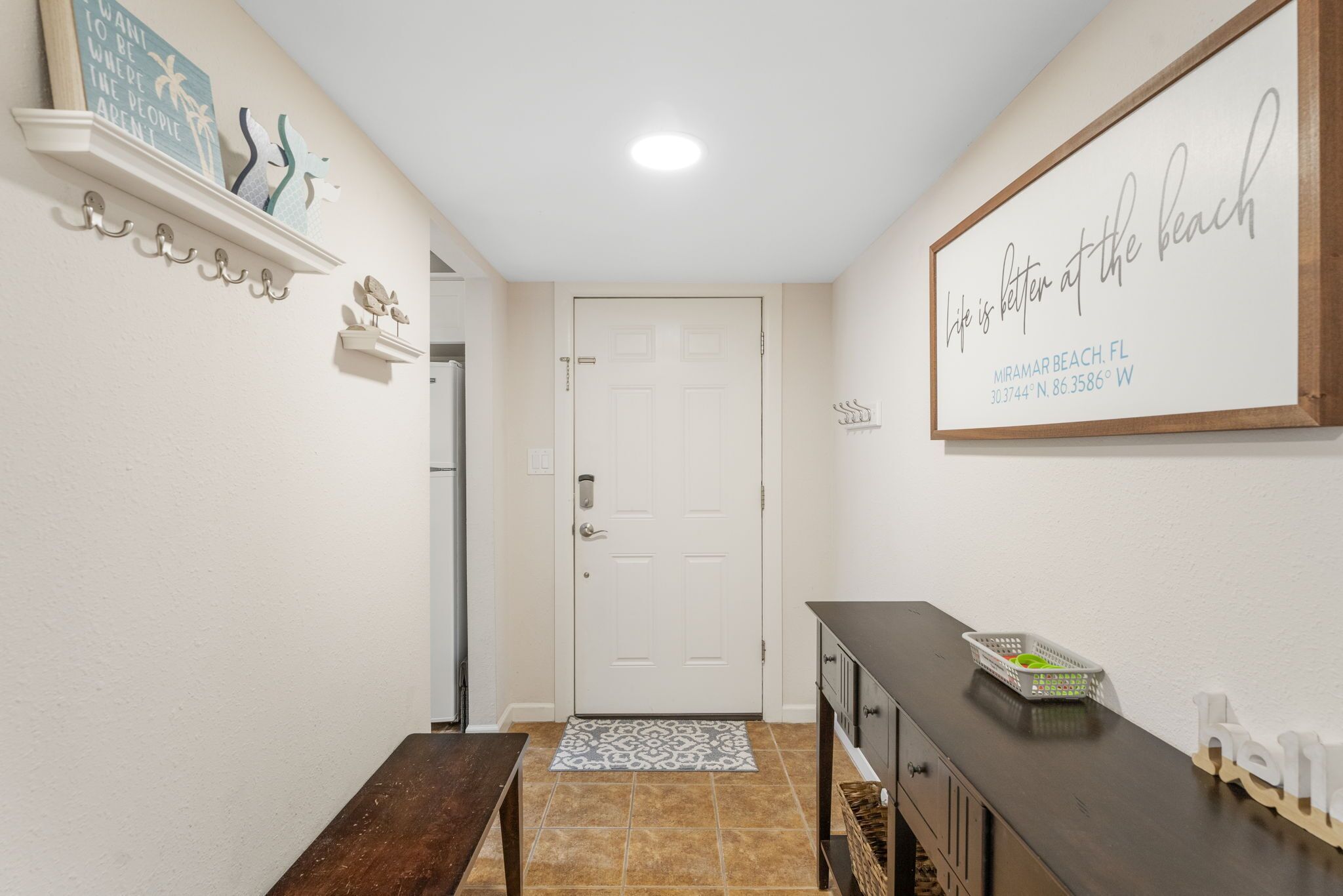 A hallway with a white door, a bench, a dark wooden table, and a wall with a sign that reads "Life is better at the beach."