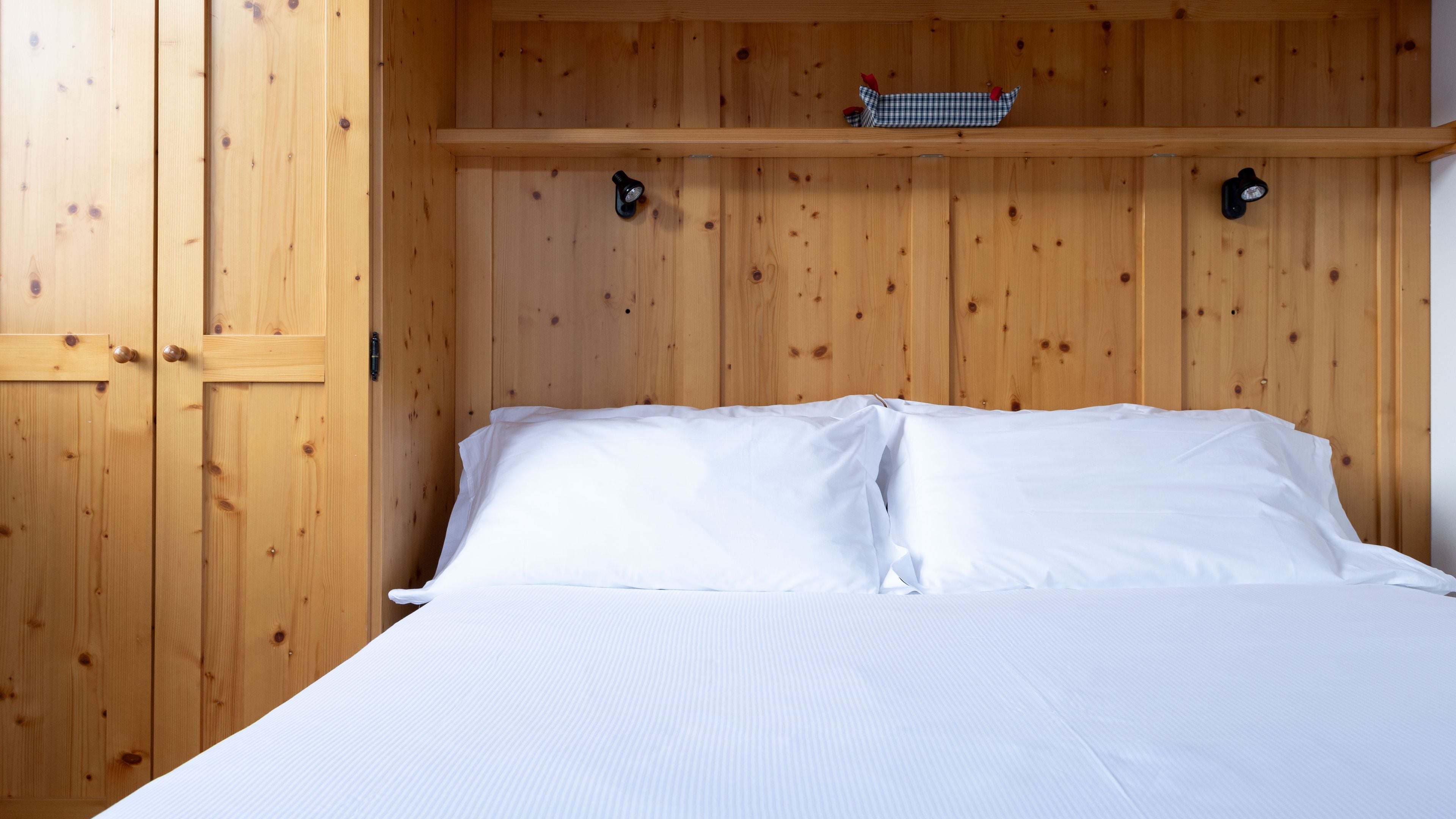 A wooden bedroom with a bed, two wall-mounted light fixtures, and a shelf above the bed.