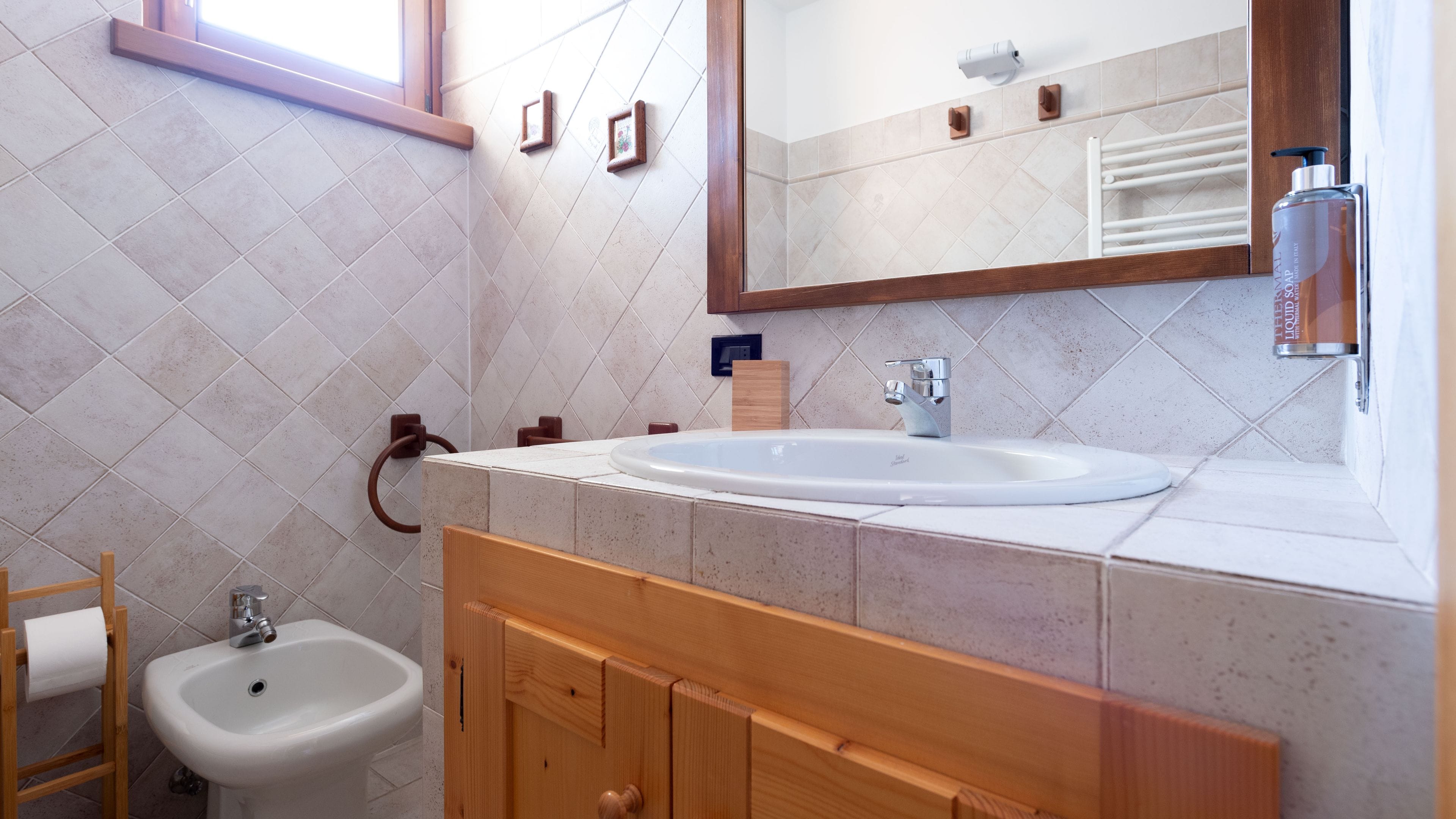 A bathroom with a white sink, wooden vanity, and a mirror. There is a towel rack, a toilet, and a window.