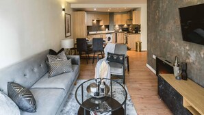 Ein modernes Wohnzimmer mit grauem Sofa, einem Couchtisch und einem an einer strukturierten Wand montierten Fernseher. Im Hintergrund ist eine Küche mit Holzschränken und einem Essbereich zu sehen.