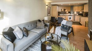 Ein modernes Wohnzimmer mit grauem Sofa, einem Couchtisch und einem Essbereich mit Stühlen. Im Hintergrund ist eine Küche mit Holzschränken und einer Waschmaschine zu sehen.