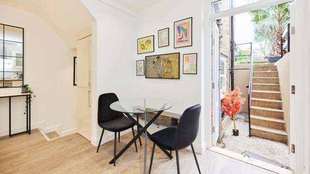 Un espace de repas moderne avec une table en verre et des chaises noires, un escalier menant à un balcon et des œuvres d’art accrochées aux murs.