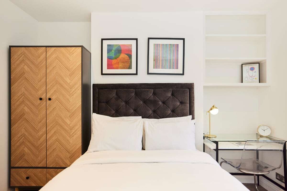 A hotel room with a bed, a wardrobe, a desk with a lamp, and two framed artworks on the wall.