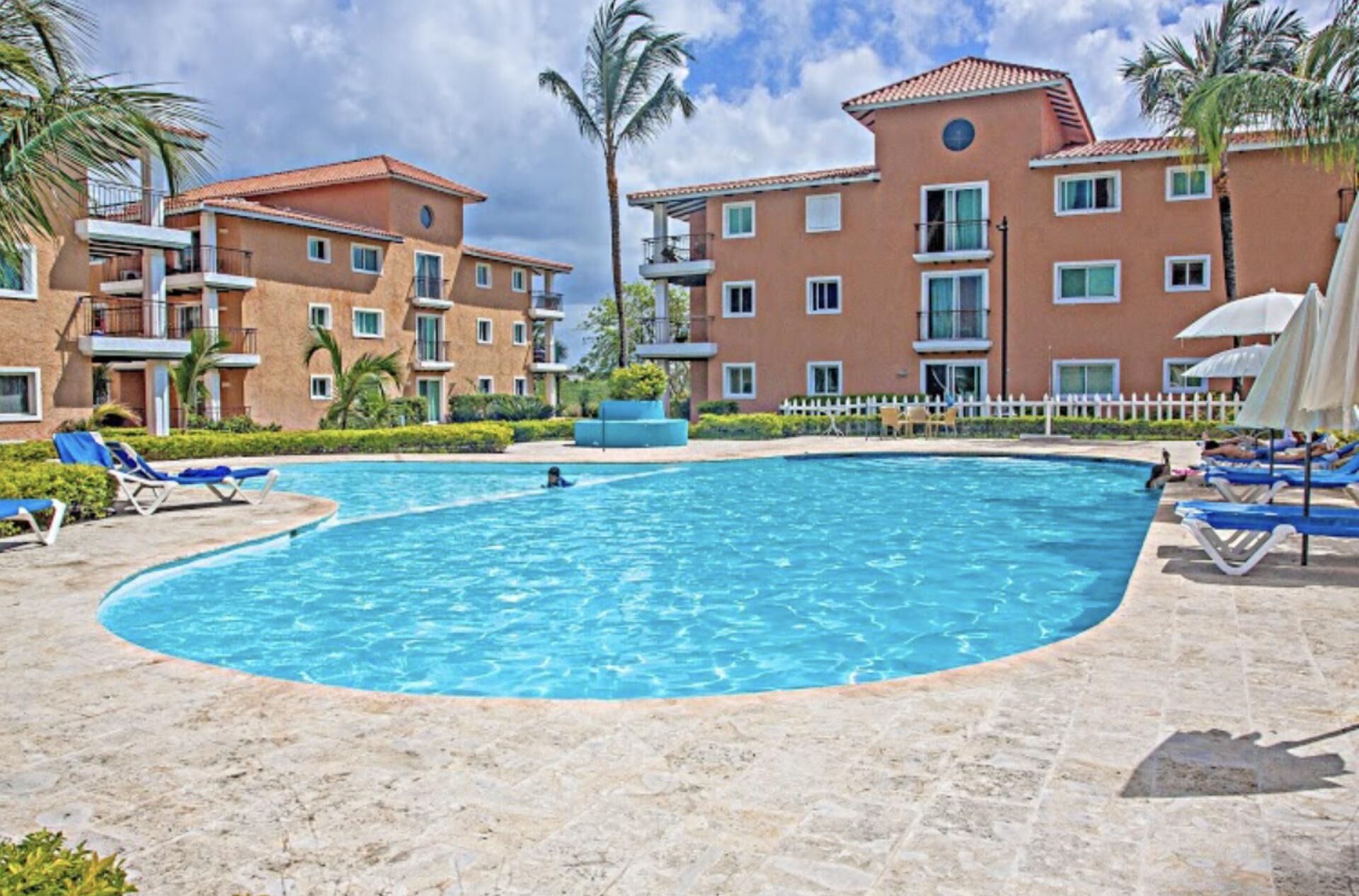 A swimming pool surrounded by lounge chairs and palm trees, with multi-story buildings in the background.