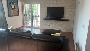 A living room with a black leather sofa, a flat-screen TV mounted on a white brick wall, and a sliding glass door leading to a balcony. - Beautiful 2-bed Apartment in Punta Cana (Punta Cana)