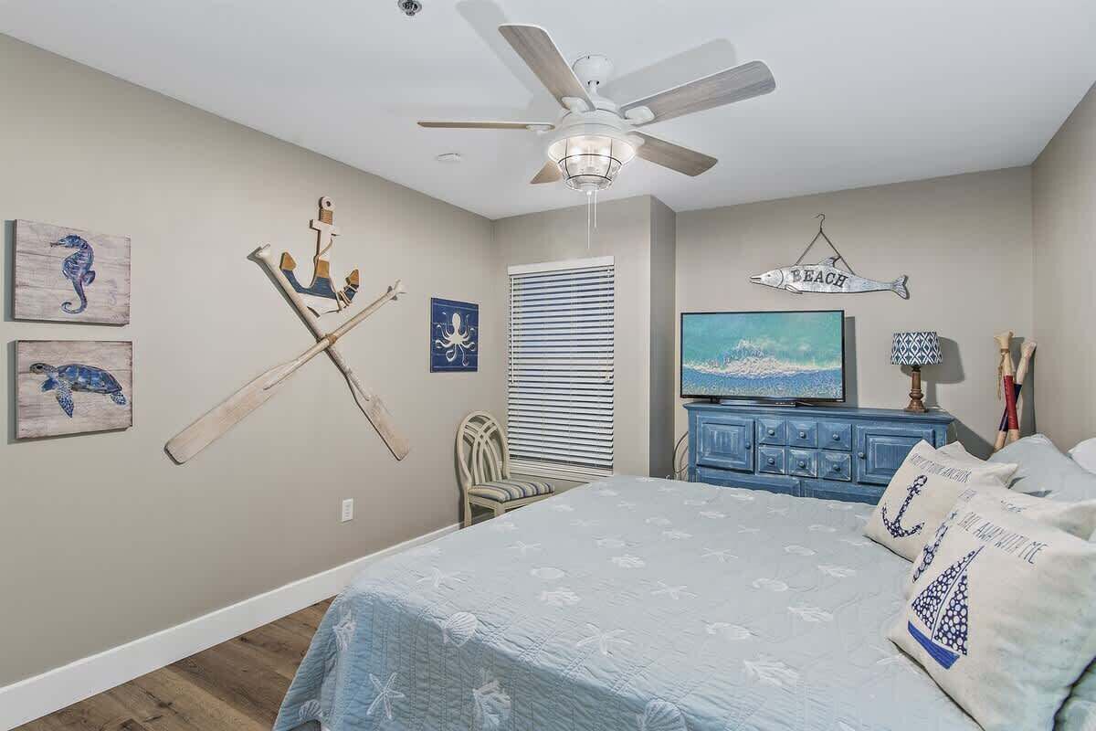 A bedroom with a bed, a ceiling fan, a TV, and wall art.