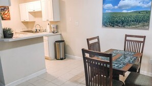 A compact kitchen with white cabinets, a stainless steel sink, and a trash can. A dining table with chairs is positioned in front of the kitchen, and a landscape painting is on the wall. - Walk to Shands & VA Hospital, UF, Pool Access (Gainesville)