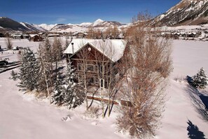 A wooden cabin with a snow-covered roof, surrounded by snow-covered trees and mountains in the background.