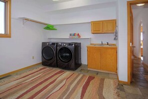 A laundry room with two front-loading washing machines, a small sink, wooden cabinets, and a striped rug on the floor.