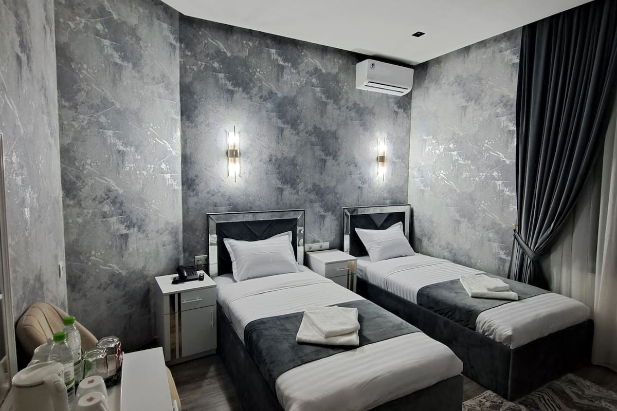 A modern hotel room with two beds, a desk, and a chair. There are wall-mounted lights and an air conditioning unit.