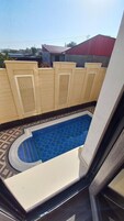 A view of a swimming pool with a tiled deck, beige walls with decorative panels, and a red-roofed building in the background.