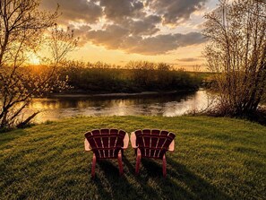 Kaksi Adirondack-tyylistä tuolia, jotka on asetettu vastakkain joen ääreen, ja taustalla näkyy auringonlasku.