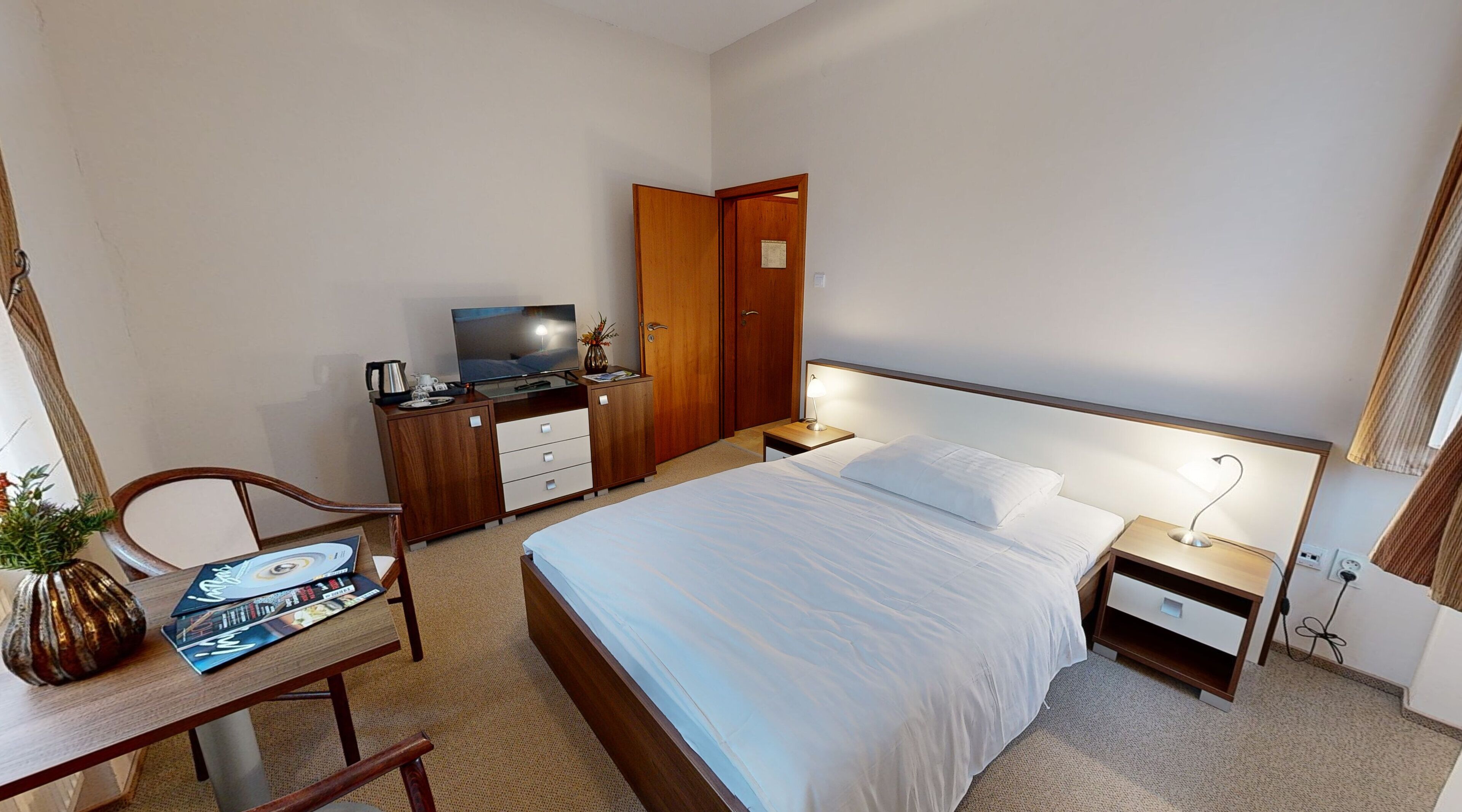 A hotel room with a bed, bedside table, TV, wooden wardrobe, desk with chair, and a small table with a vase.
