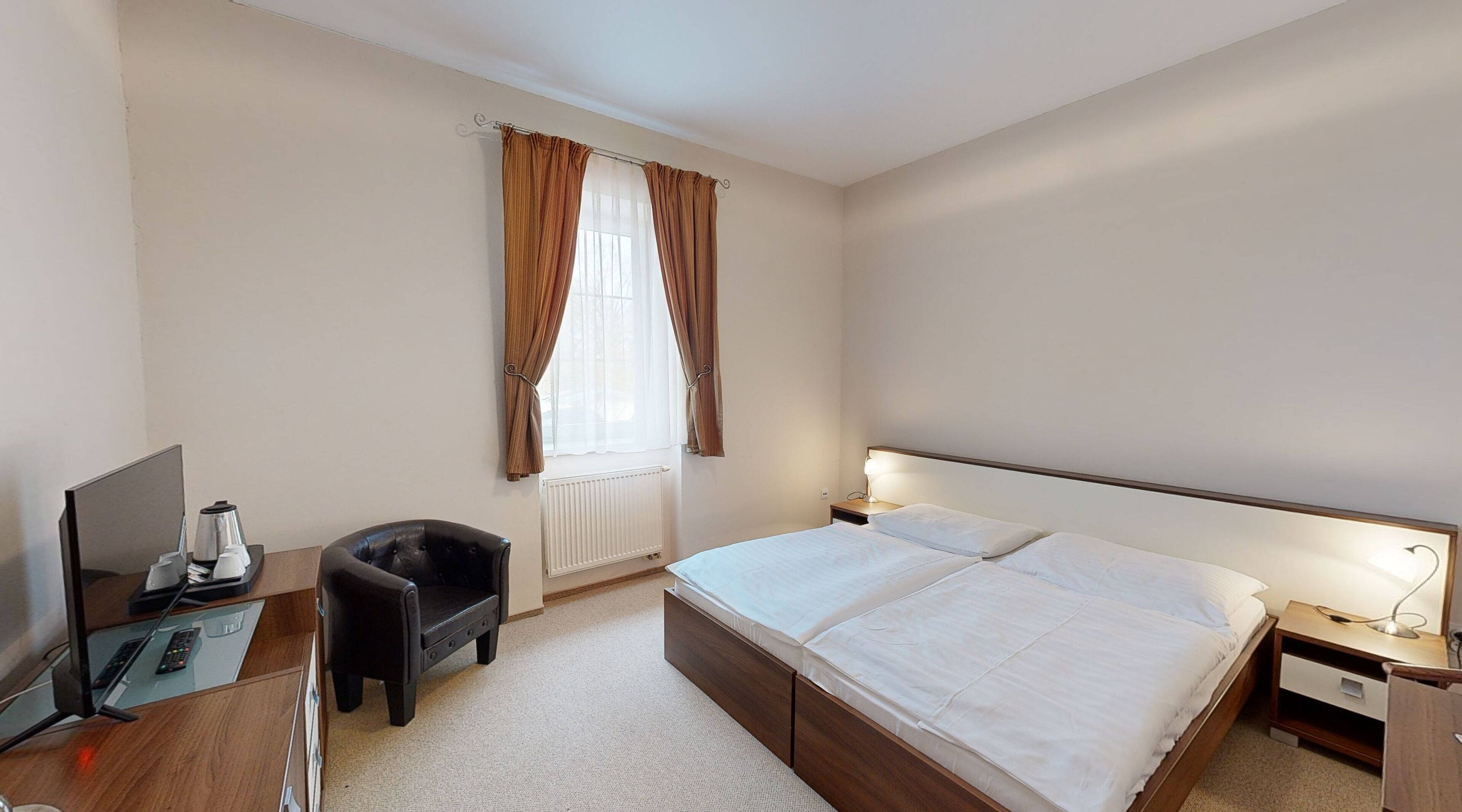 A hotel room with a bed, bedside table, lamp, chair, desk with a TV and remote controls, and a window with curtains.