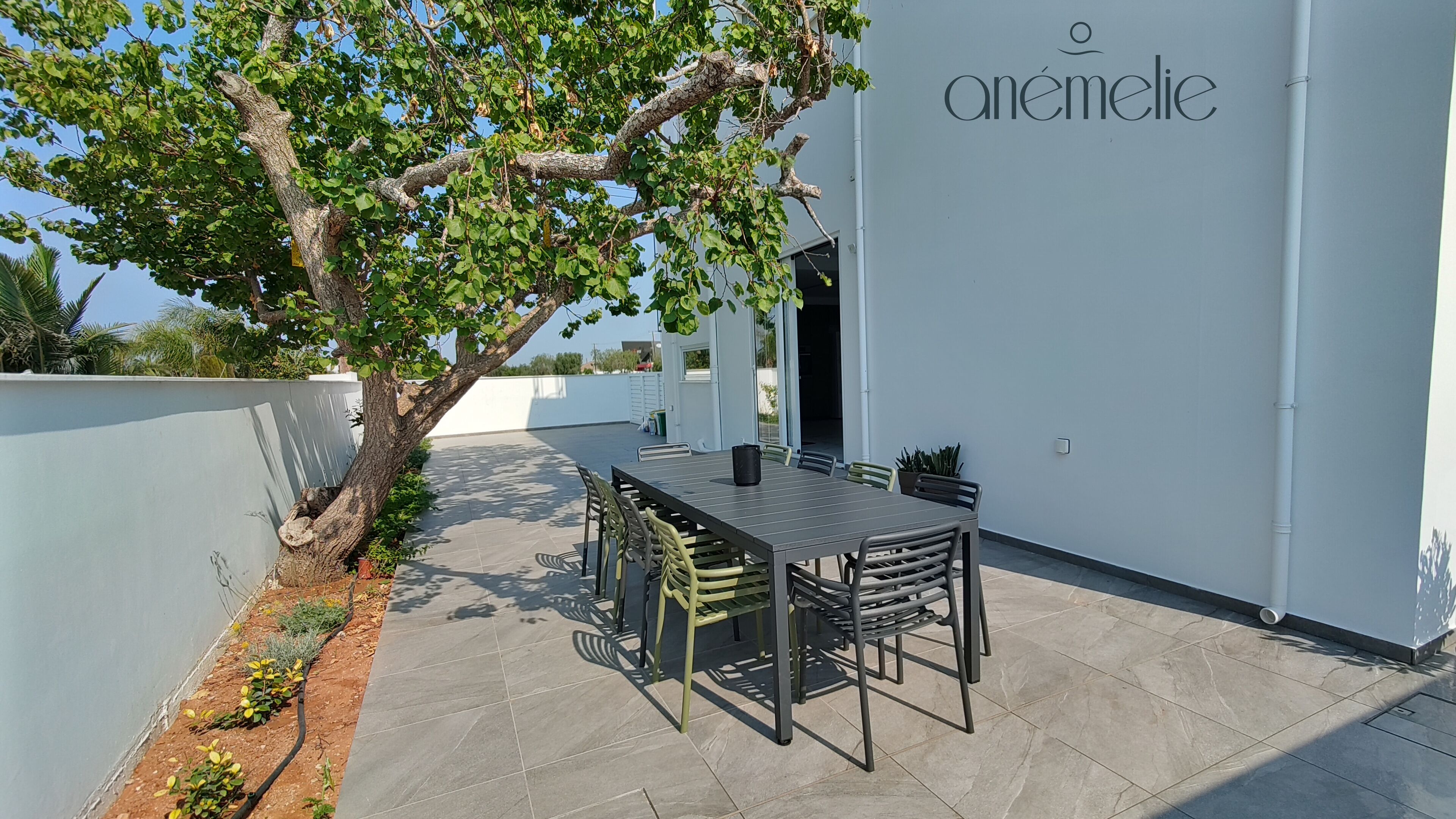 Eine Terrasse im Freien mit Tisch und Stühlen, einem Baum und einem Gebäude mit der Aufschrift 'anémelie' an der Wand.