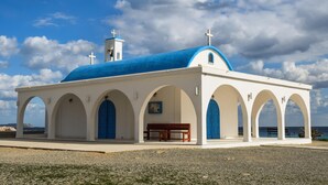 A white church with a blue roof and arches, a bench, and a view of the sea.