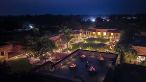 A well-lit outdoor dining area with tables and chairs, surrounded by lush greenery and a stone wall.