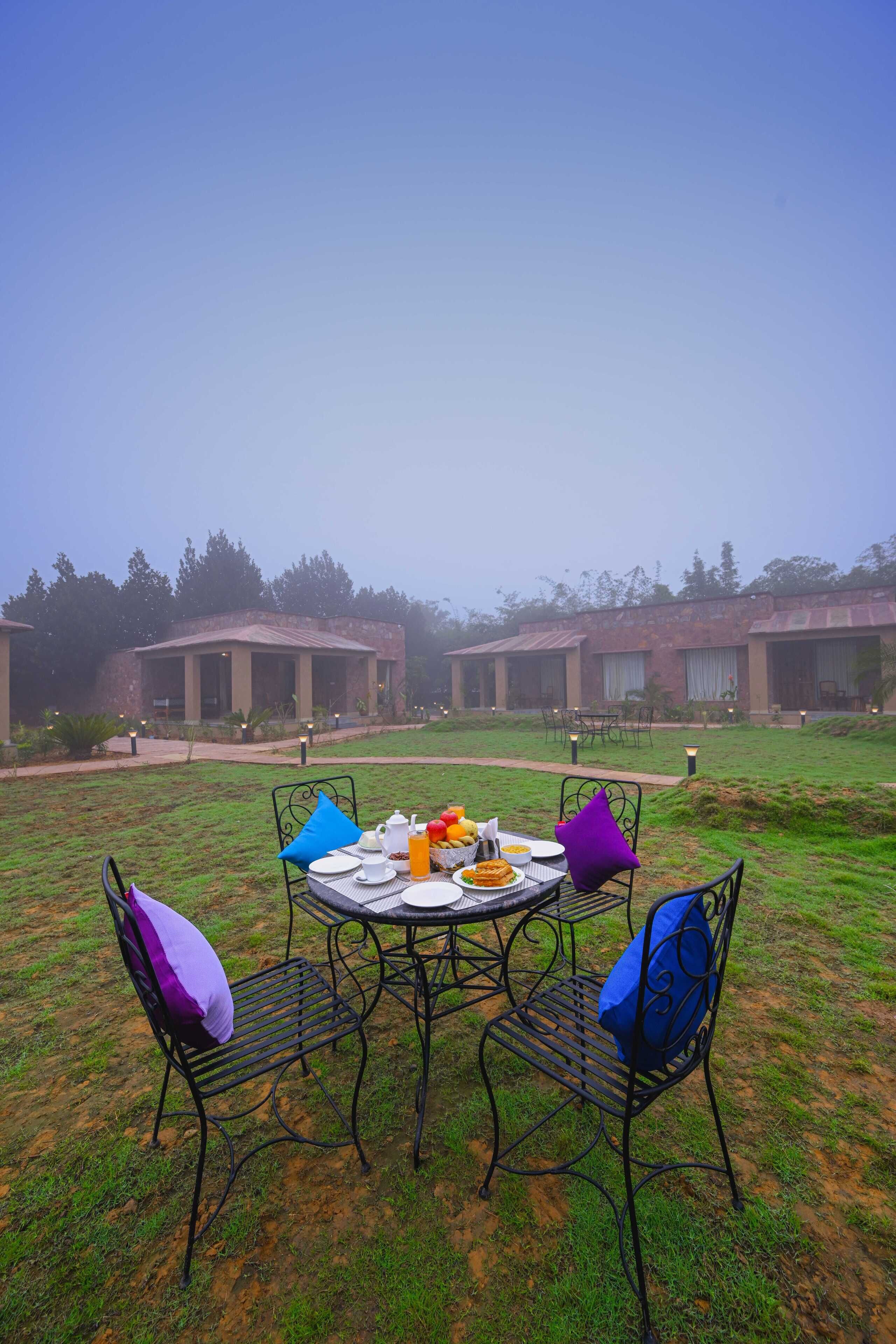 An outdoor dining area with a round table and metal chairs, set against a backdrop of buildings and a clear sky.