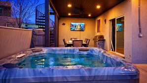 A hot tub with a wooden deck, a dining area with chairs and a table, and a TV mounted on the wall. - Desert Hideaway: 3-Bedroom Home with Year-Round Private Pool & Hot Tub (Moab)