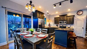 A modern kitchen with dark cabinetry, a central island, and a dining area with chairs and a table set for a meal. - Exclusive Retreats Desert Hideaway 3 Bedroom all Suite home with Private pool #2 (Moab)