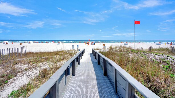 Une promenade en bois menant à une plage où flotte un drapeau rouge, bordée de dunes de sable et fréquentée par des baigneurs.
