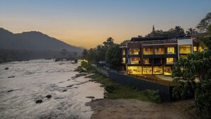Un hotel moderno con varios pisos, un río al frente y montañas en el fondo.
