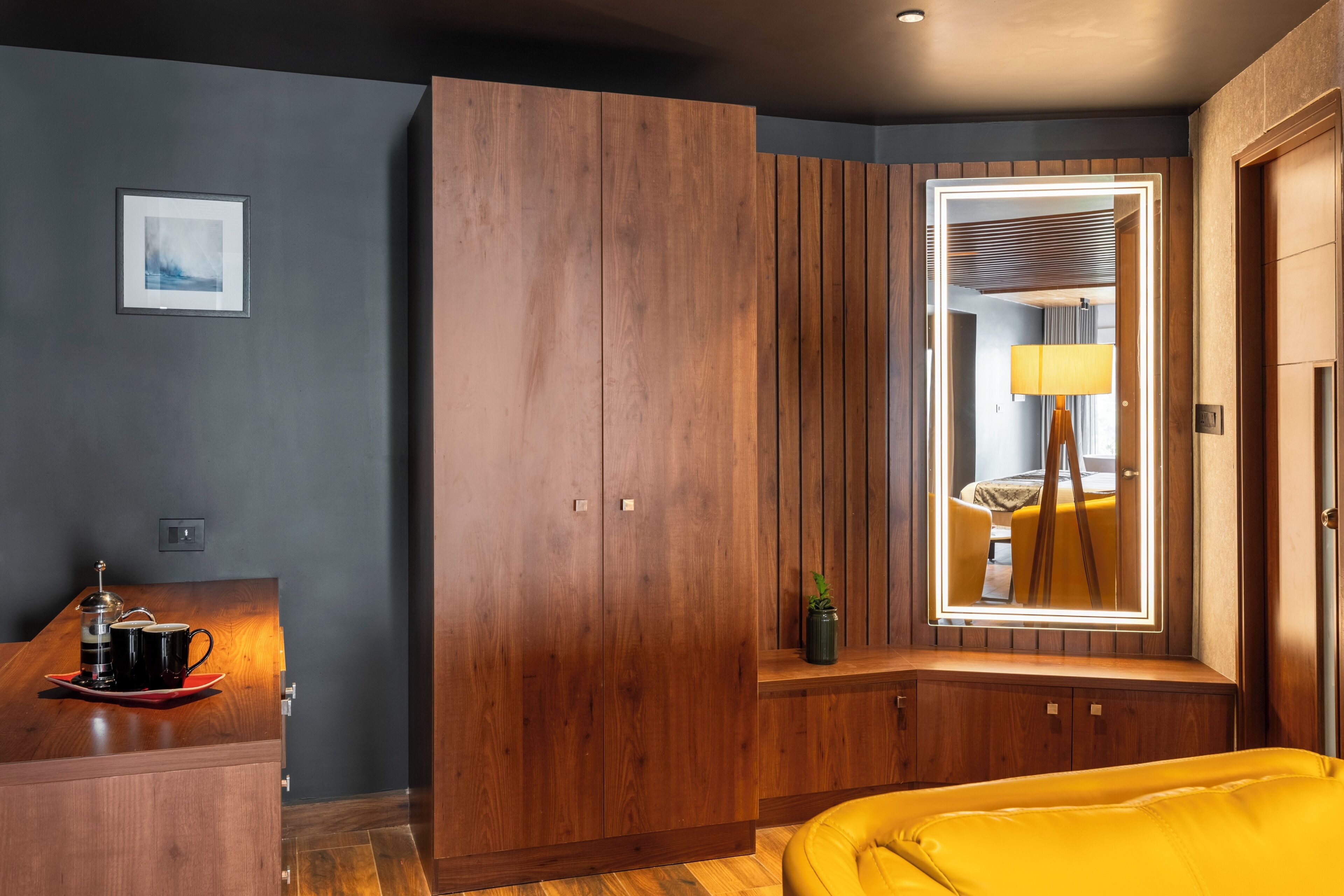 Una habitación de hotel moderna con un armario de madera, una mesita de noche con cafetera y un espejo grande con marco iluminado.