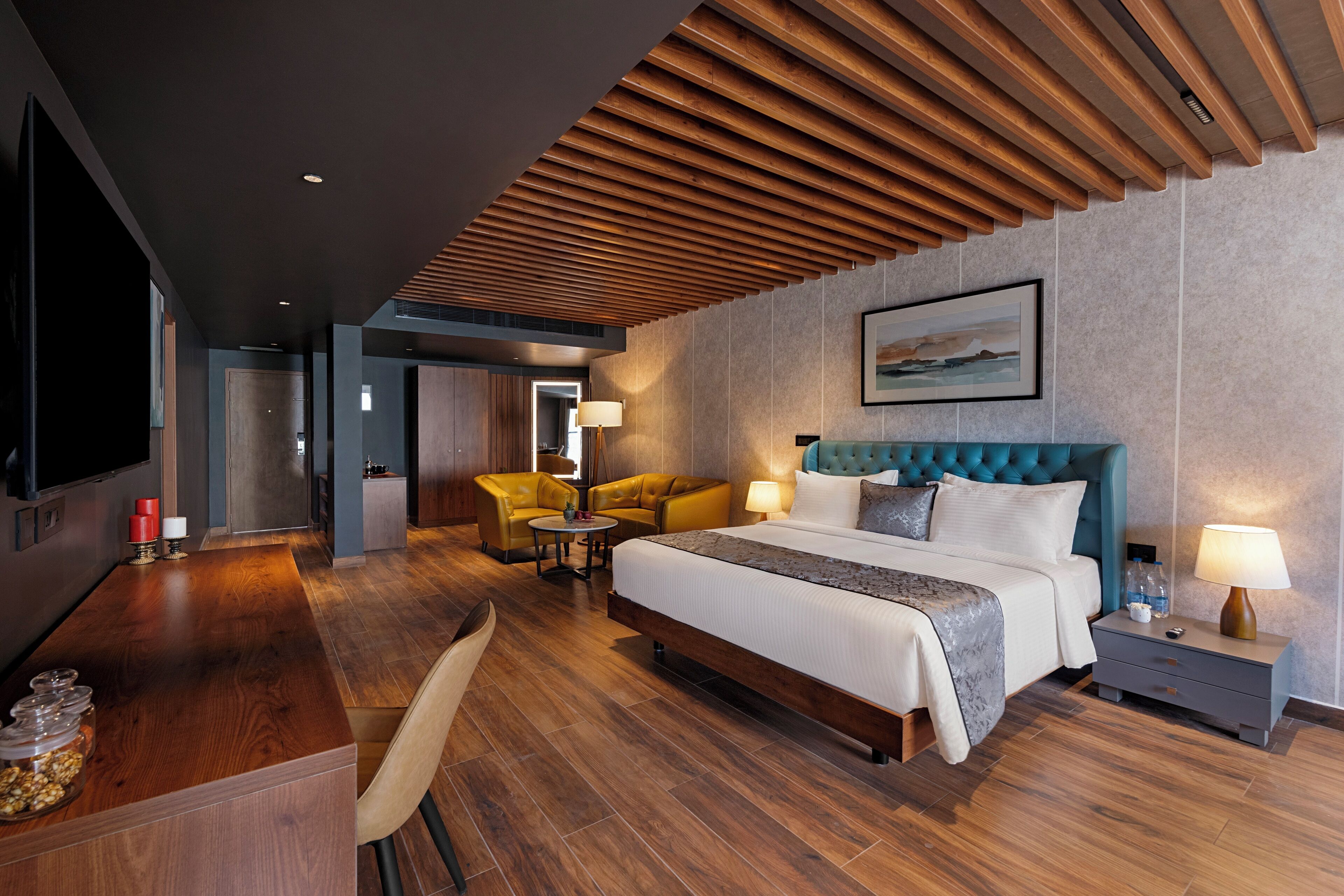 Quarto de hotel moderno com uma cama grande, teto de madeira, televisão, escrivaninha com cadeira e uma pequena mesa com cadeiras.