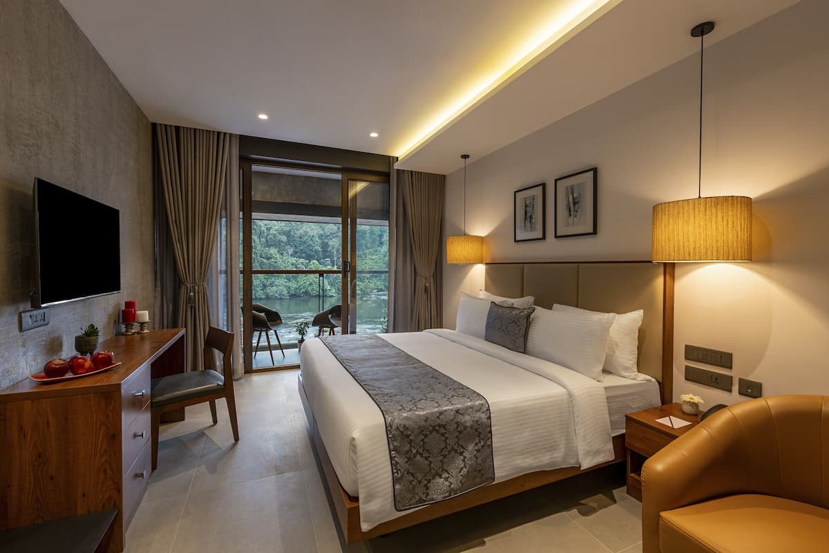 A modern hotel room with a large bed, a desk with a flat-screen TV, a dining table, and a view of a lake.