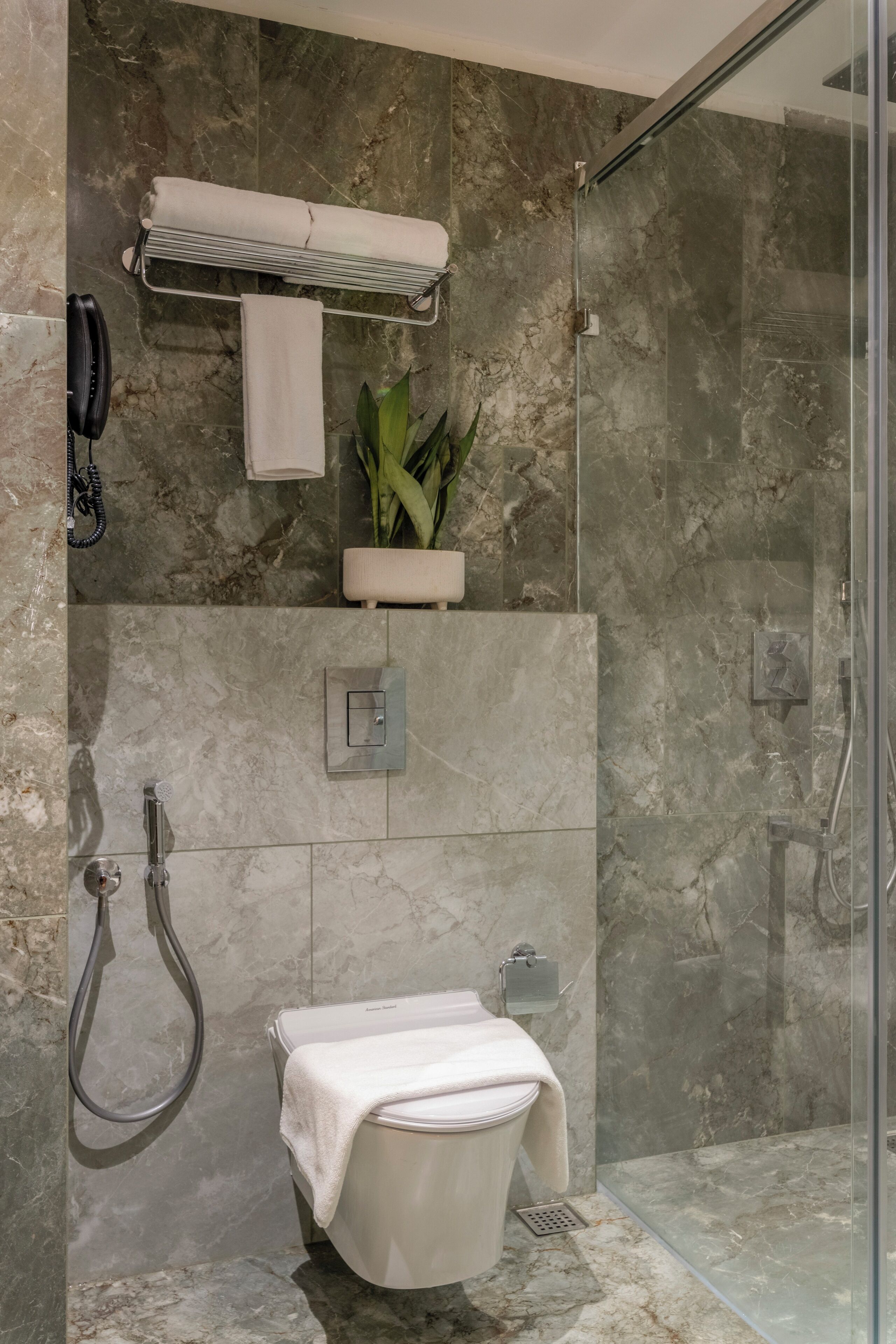 A modern bathroom with a glass-enclosed shower, a toilet with a towel, a towel rack, a plant, and a phone on the wall.
