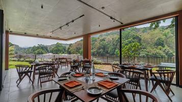 Un ristorante con posti a sedere all'aperto, tavoli apparecchiati con piatti e posate, e vista su un paesaggio boschivo.