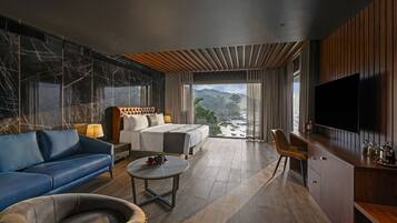 A modern hotel room with a large bed, a sofa, a coffee table, and a TV. The room has a view of a mountainous landscape.