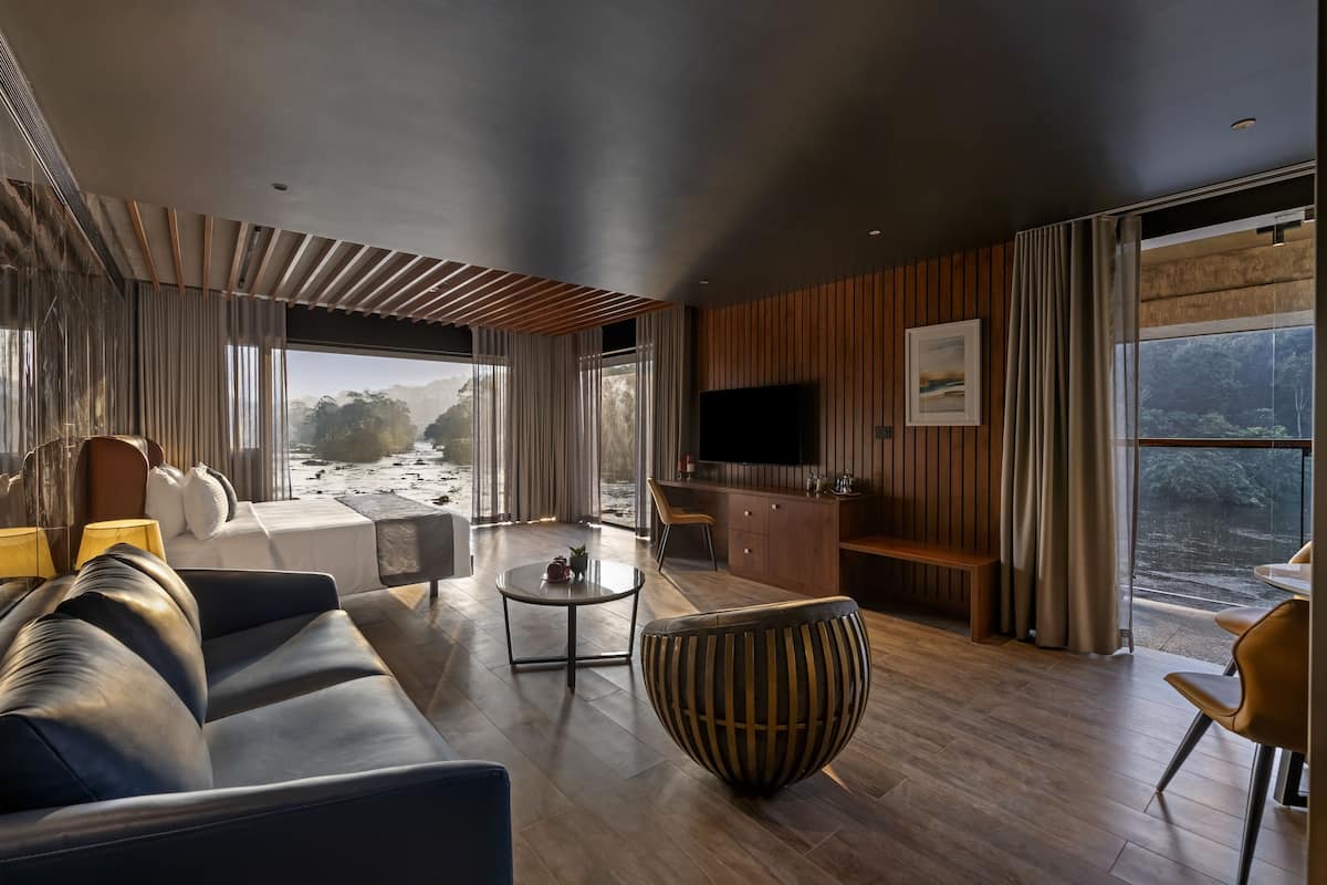 A modern living room with a large sofa, a coffee table, and a TV. There is a wooden panel wall and a large window with curtains.