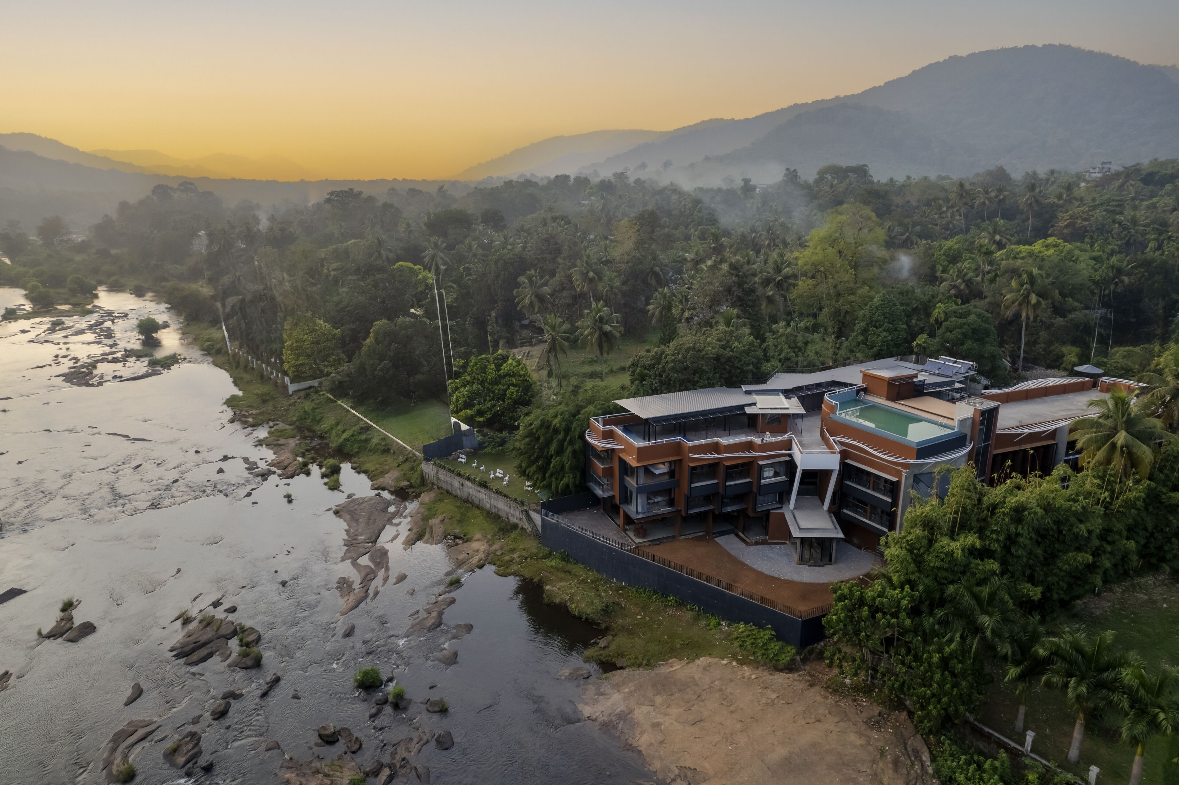 A modern multi-story building with a curved roof, surrounded by lush greenery and a river.