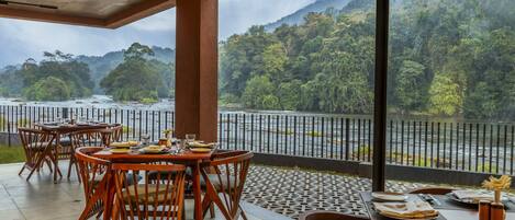 An outdoor dining area with tables and chairs, overlooking a river and forested hills.