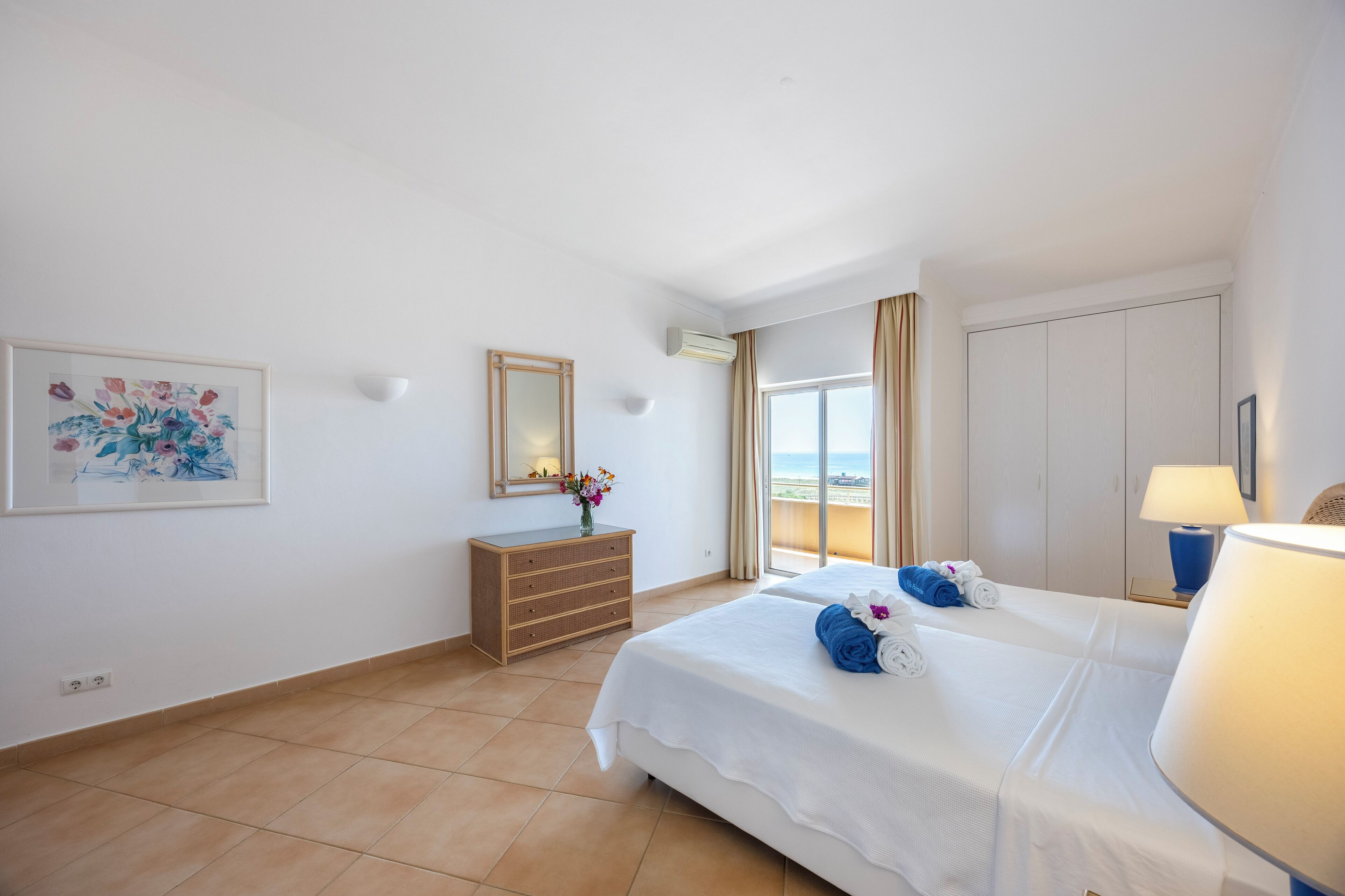 A hotel room with a bed, a dresser, a mirror, a painting, and a view of the sea.