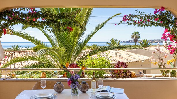 Un tavolo apparecchiato per due con vista mare, circondato da piante tropicali e un pergolato fiorito.