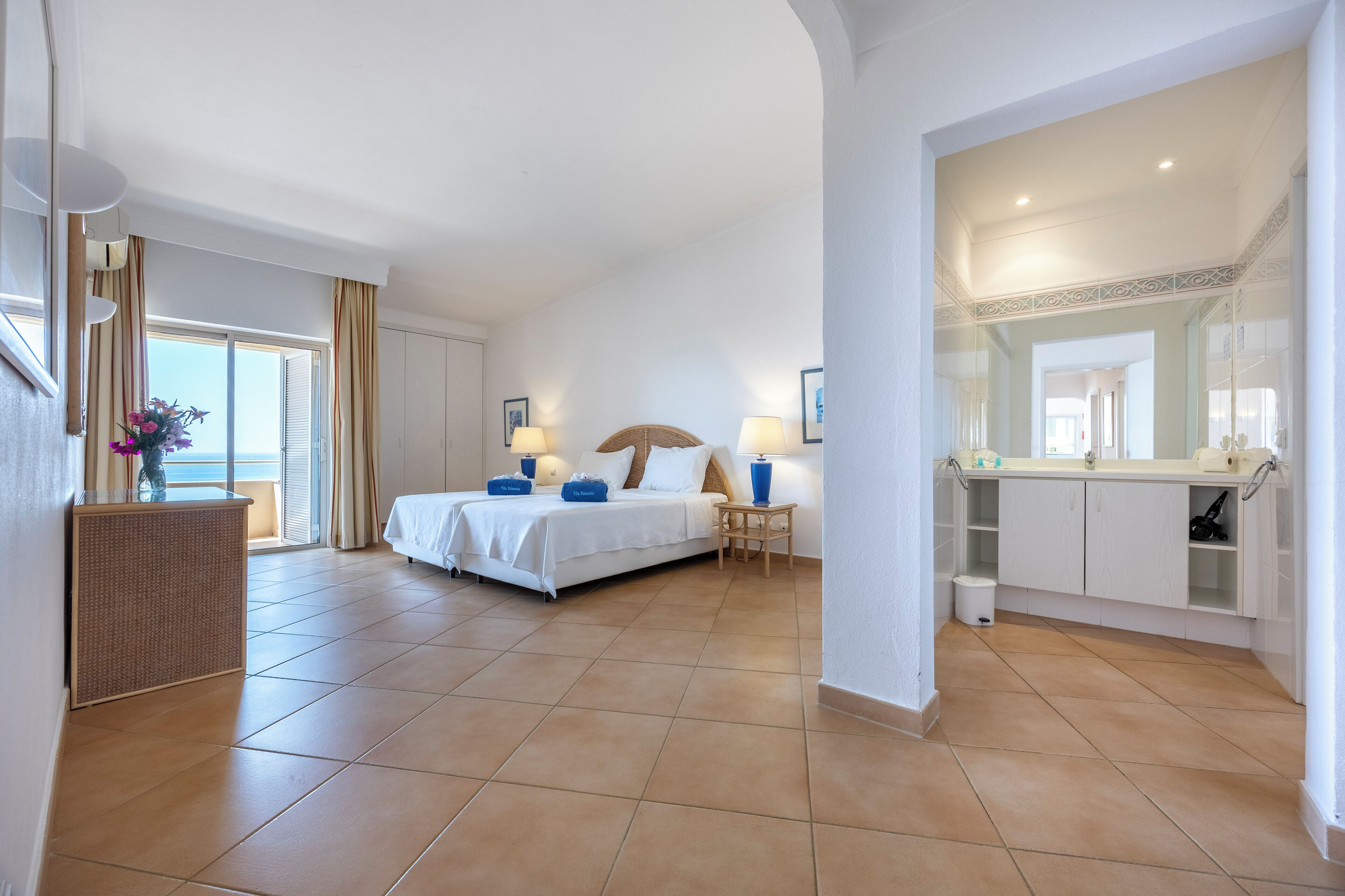 Une chambre d’hôtel comprenant un lit, des tables de chevet, une petite table avec un vase et une salle de bain avec un miroir et des placards.