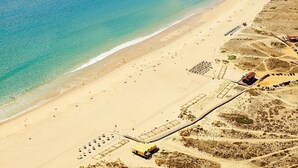 Vista aerea di una spiaggia con un lungo molo che si protende nell'acqua, una spiaggia sabbiosa e alcuni edifici sullo sfondo.