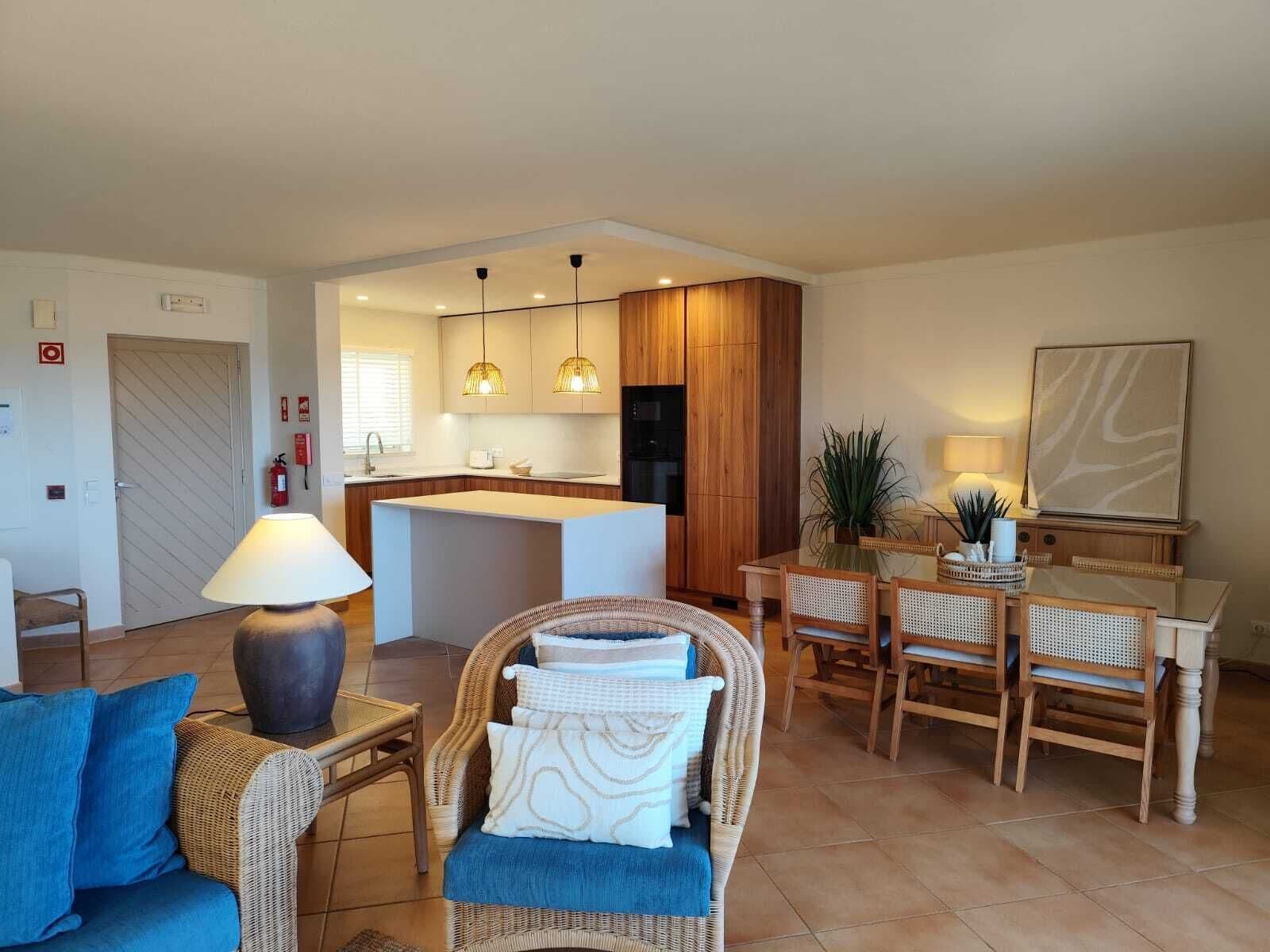 A modern living room with a sofa, a dining table with chairs, a kitchen area with wooden cabinets, and pendant lighting.