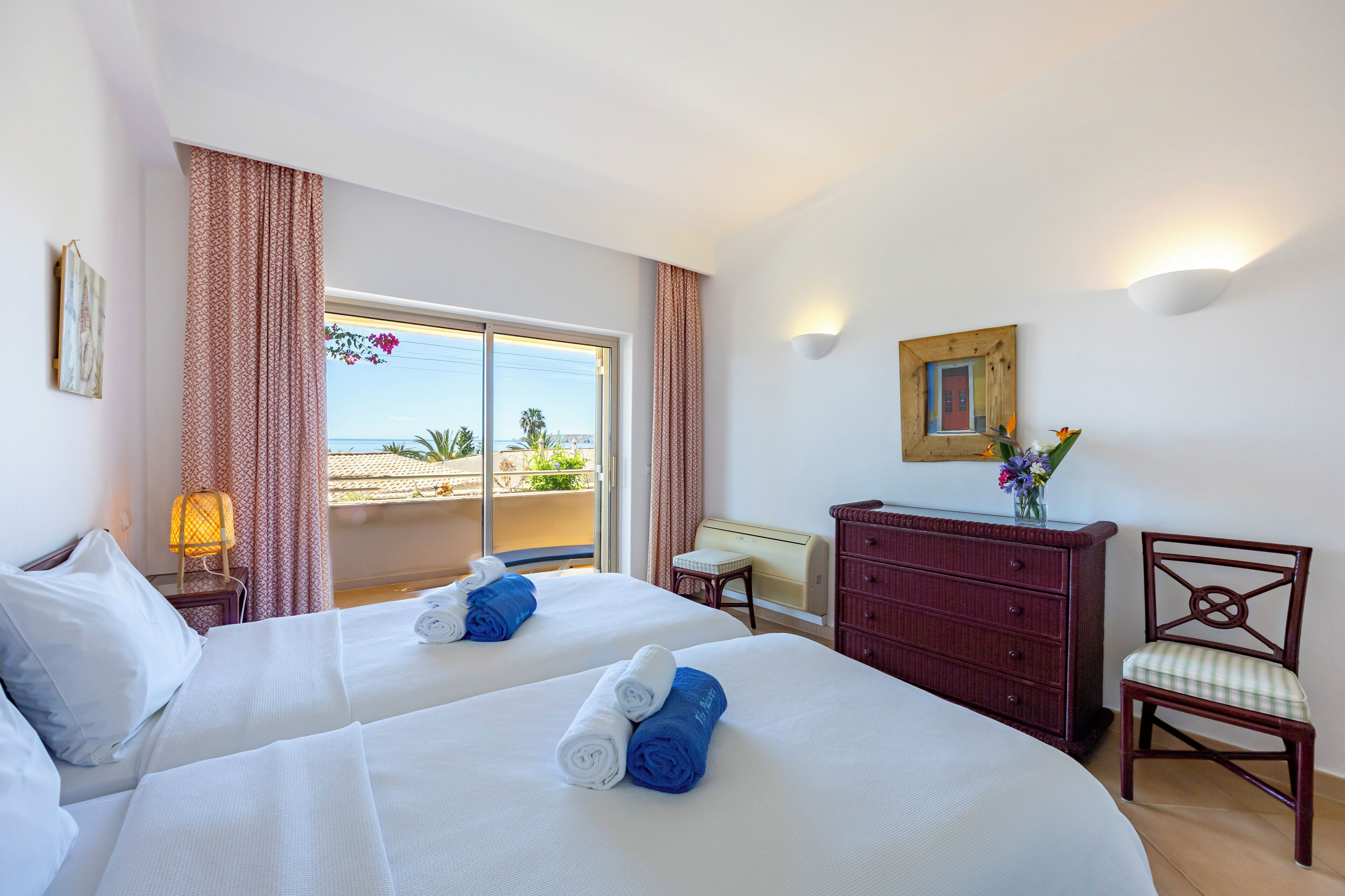 A hotel room with two beds, a dresser, a chair, a lamp, and a view of the outdoors.