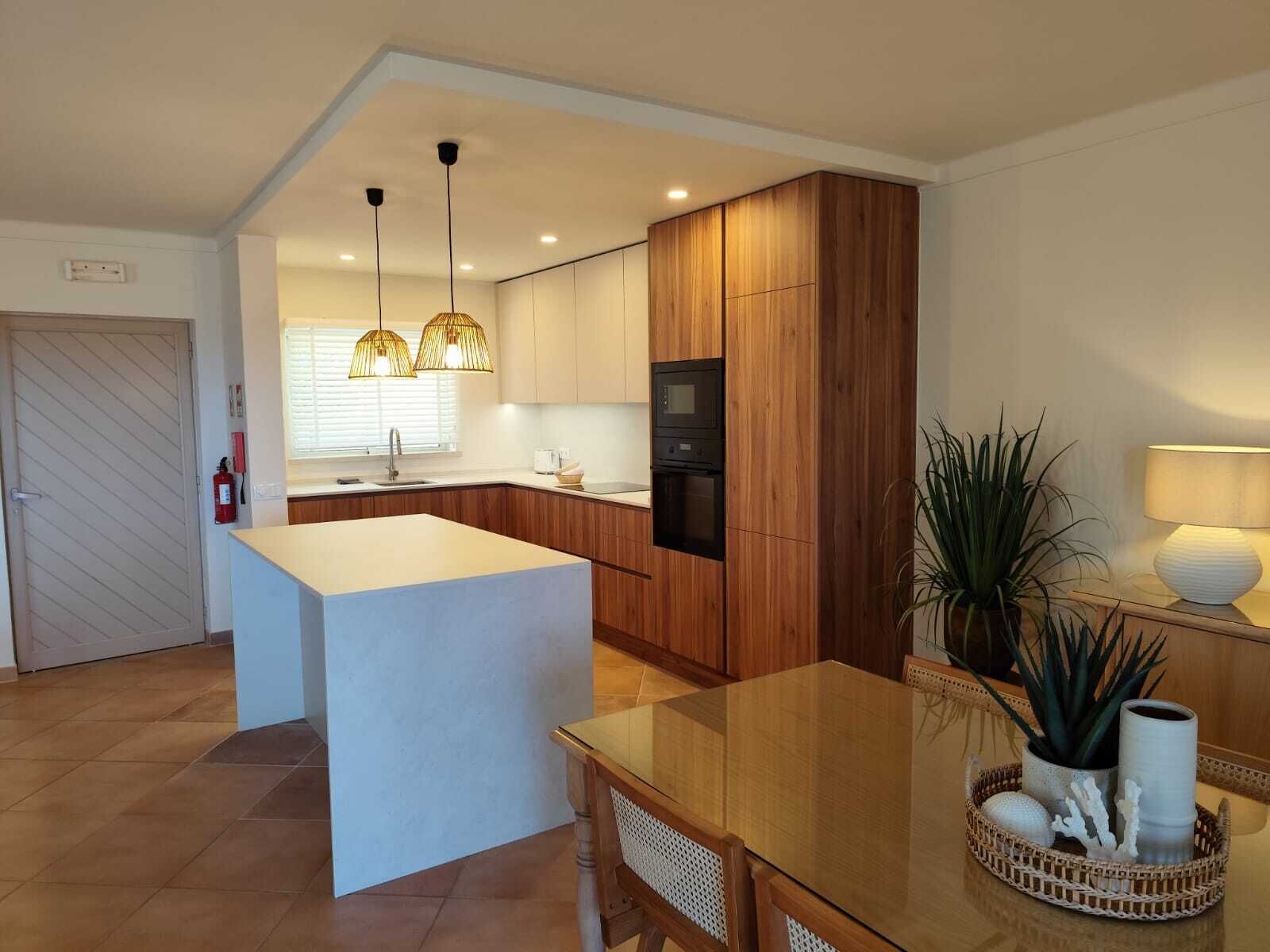 A modern kitchen with wooden cabinets, a central island, pendant lighting, and a dining area with a table and chairs.