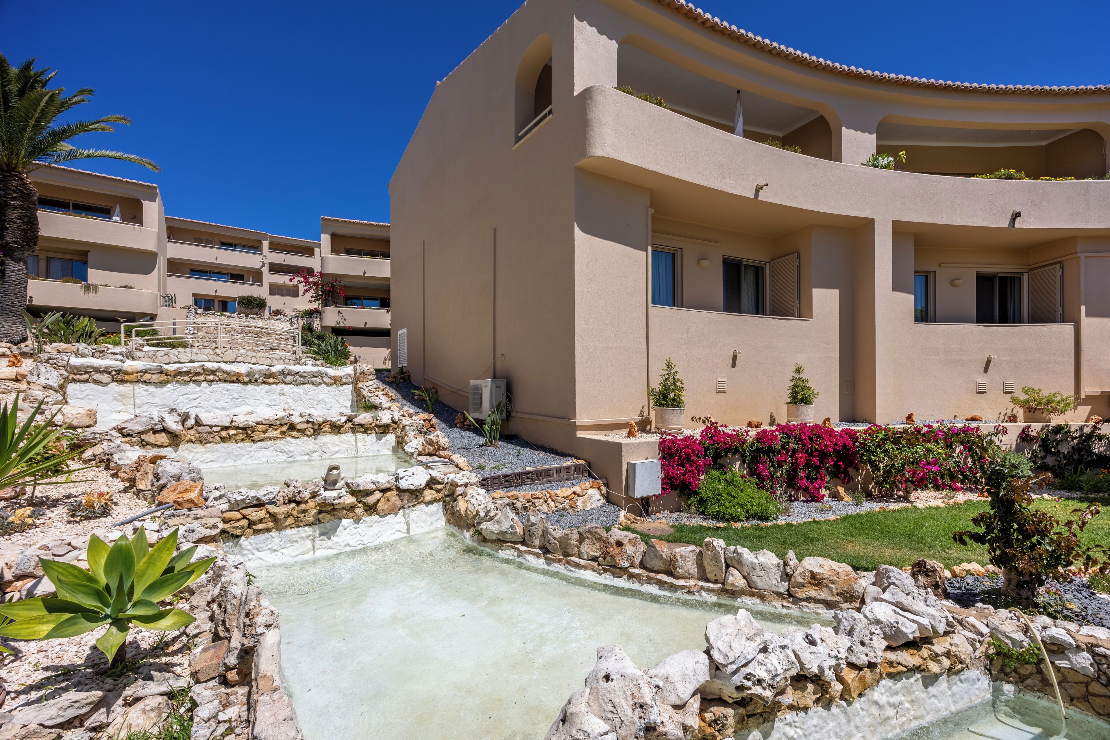 A hotel with multiple balconies, a pool area with a stone pathway, and a garden with pink flowers.