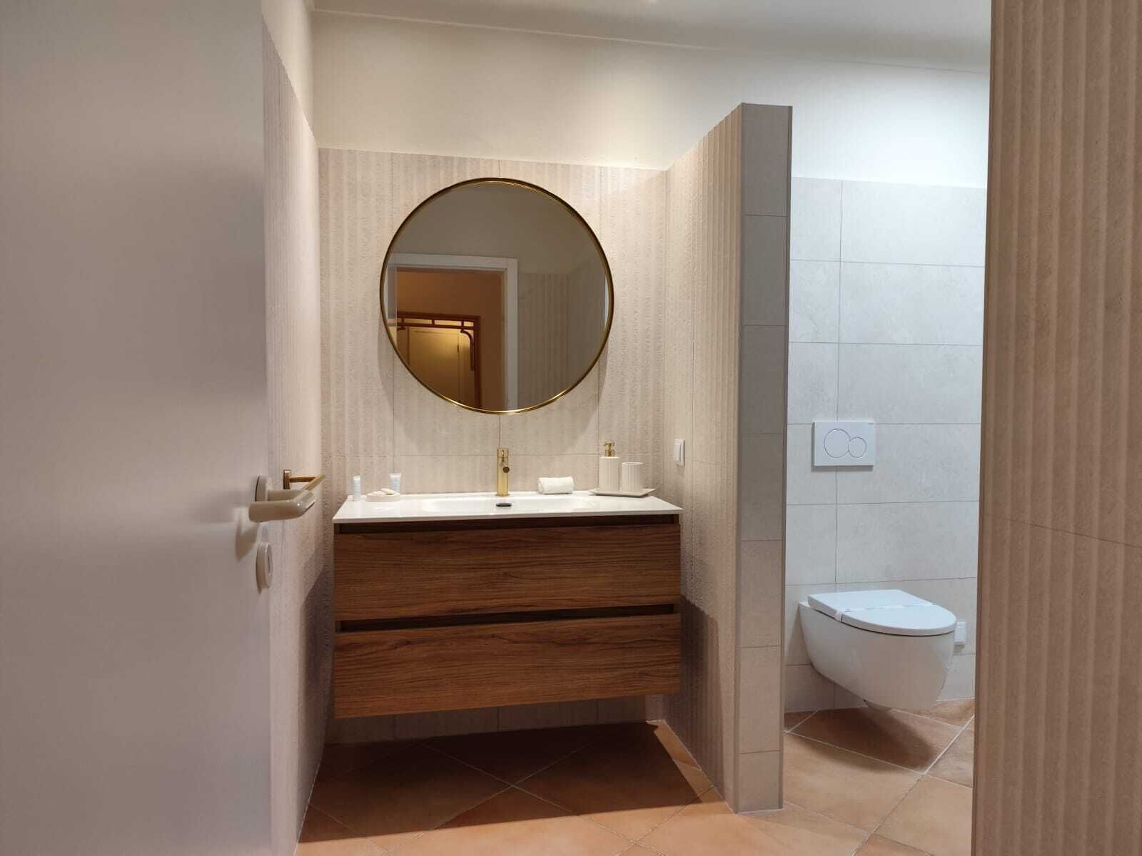 Et moderne badeværelse med et rundt spejl, trækommode, hvid vask og toilet.