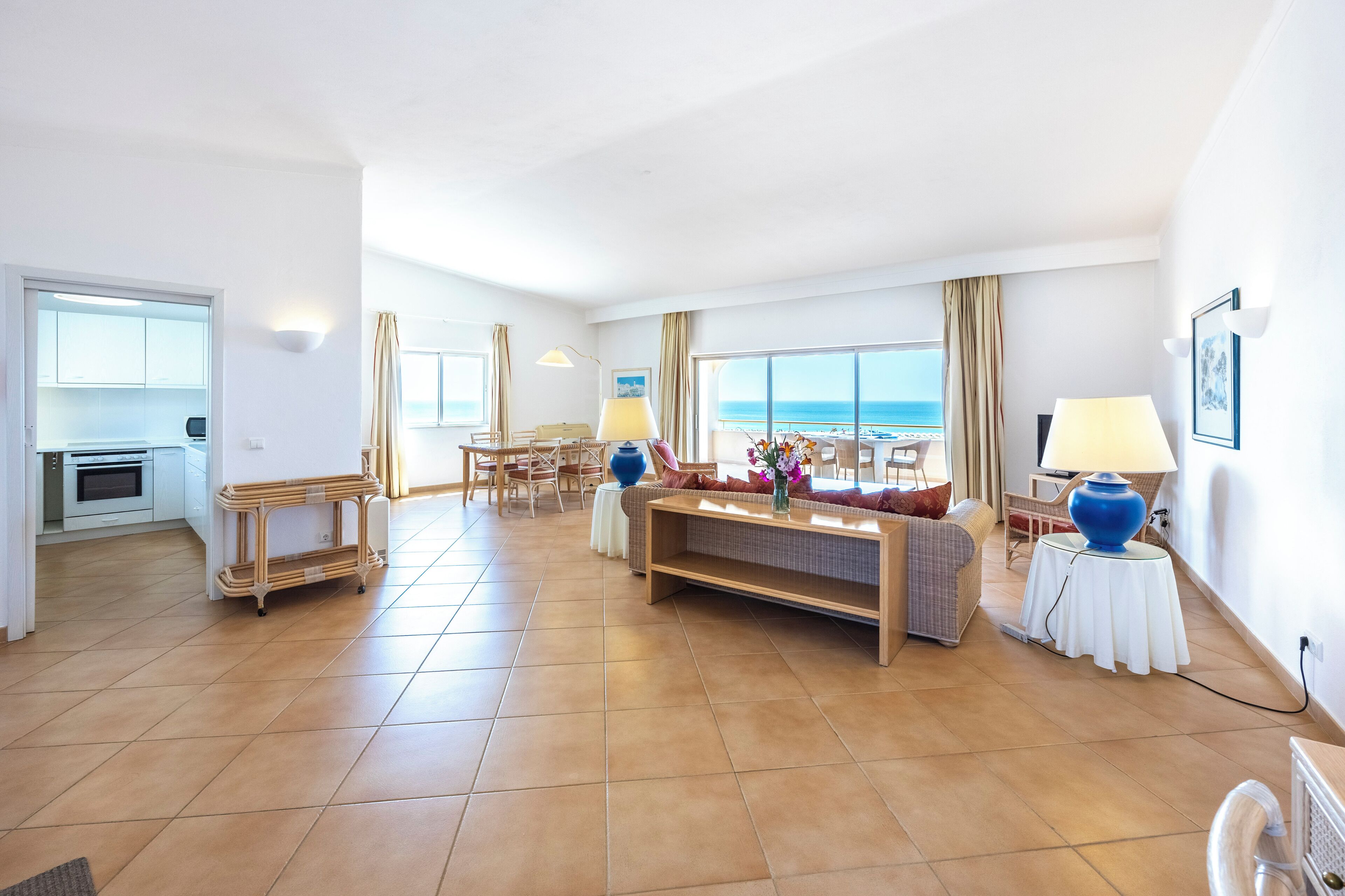 A spacious living area with a sofa, dining table, and chairs, a kitchen in the background, and a large window offering a view of the sea.